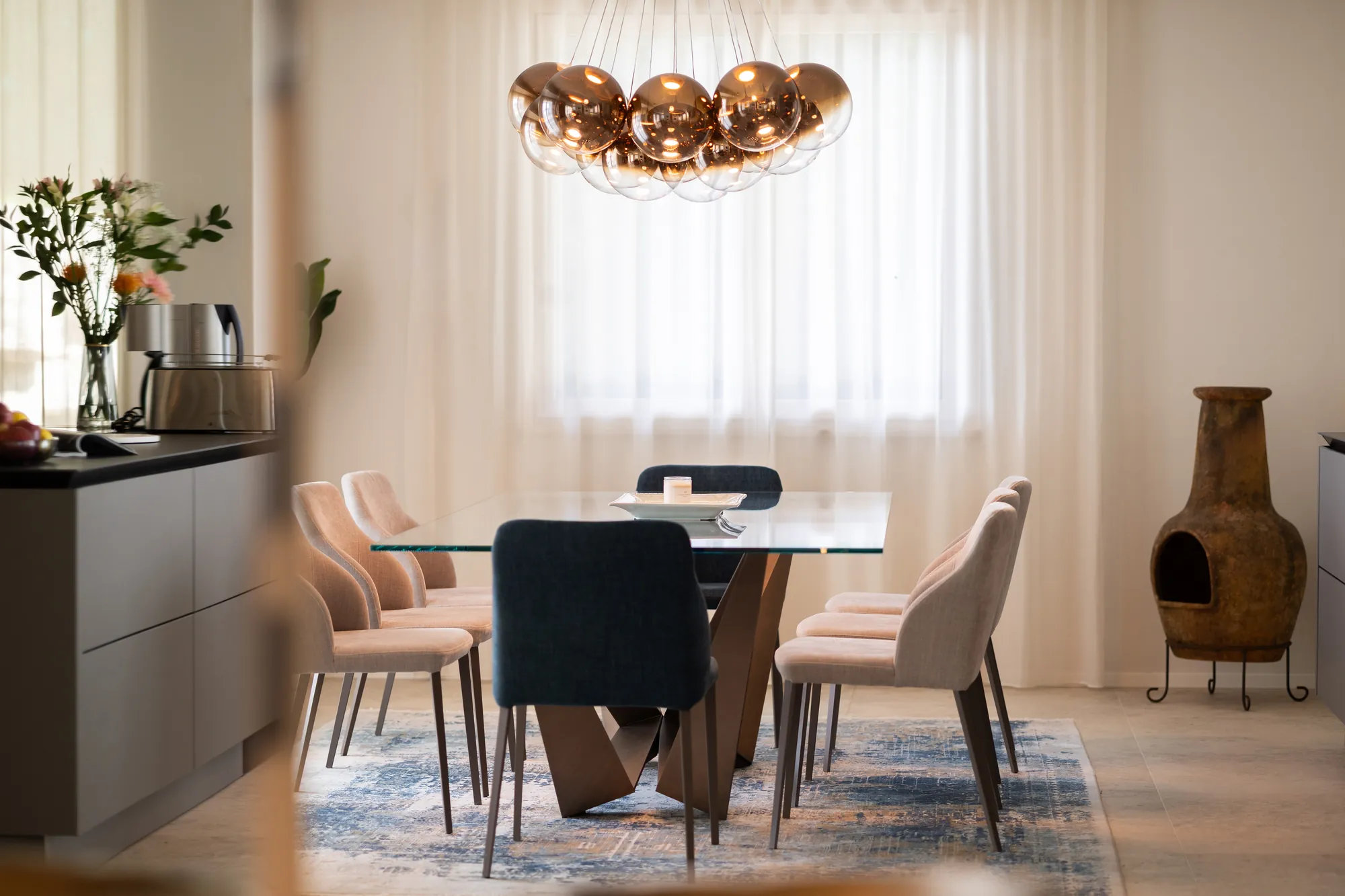 Glass dining table and upholstered chairs beneath a copper sphere chandelier